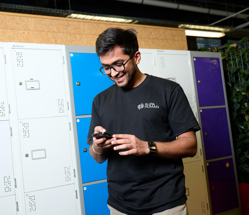 Young man with glasses smiling while using smartphone in front of colorful lockers in a modern office setting. He is wearing a black "Supa Human" branded t-shirt, standing casually, suggesting a relaxed and innovative tech work environment. Keywords: office lockers, tech startup, employee using phone, casual dress code, Supa Human brand, modern workplace.