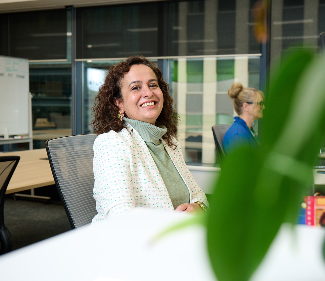 Smiling woman in a white and green blazer seated at a desk in a bright, modern office with large windows and glass walls. A blurred green plant in the foreground adds depth, while another employee works at a computer in the background. Keywords: office portrait, professional woman, natural lighting, contemporary workspace, employee at desk, Supa Human office interior.