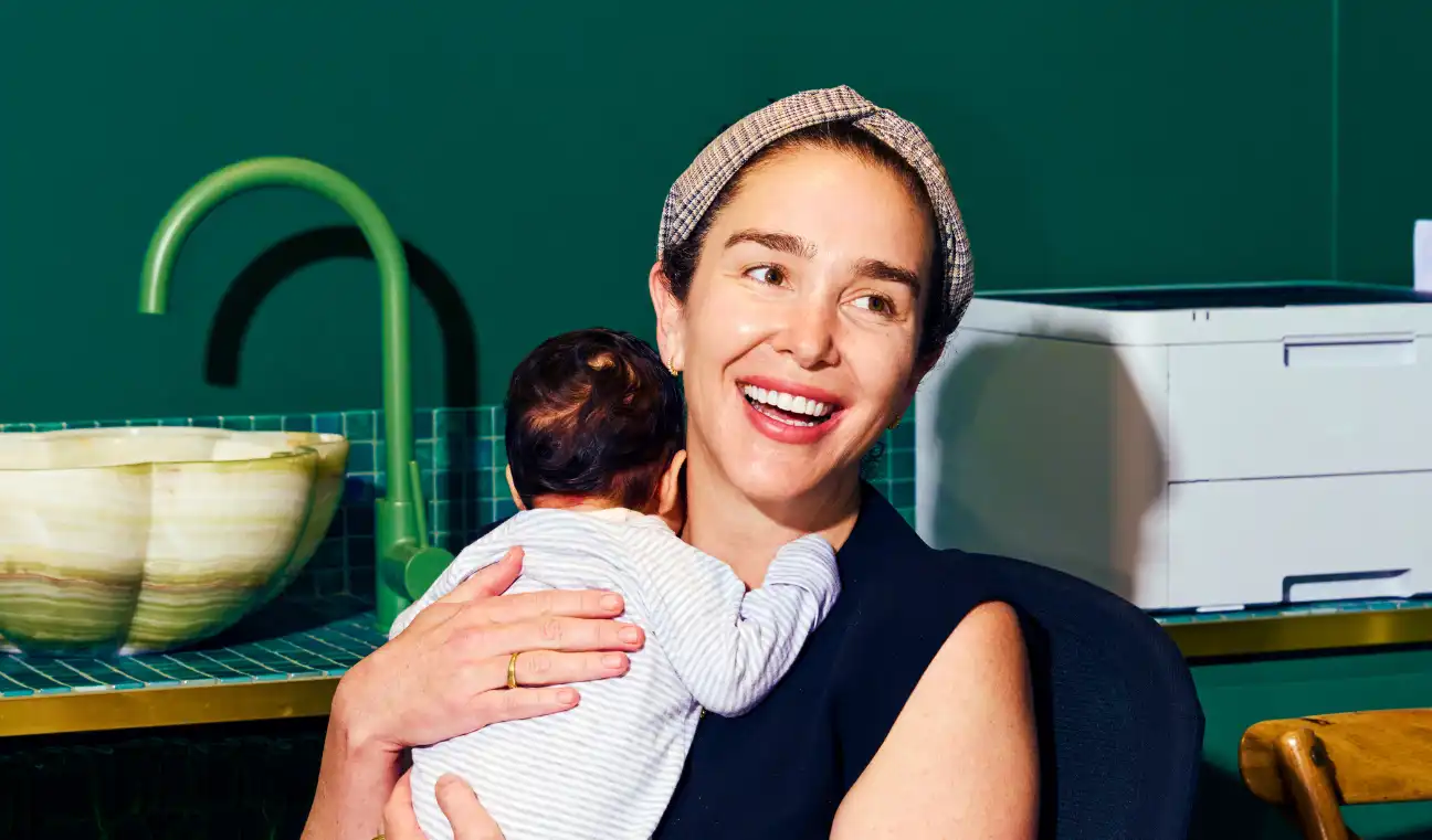 Smiling woman holding a baby against a teal wall with a green faucet and beige sink. She wears a headband and black top, exuding warmth and joy.