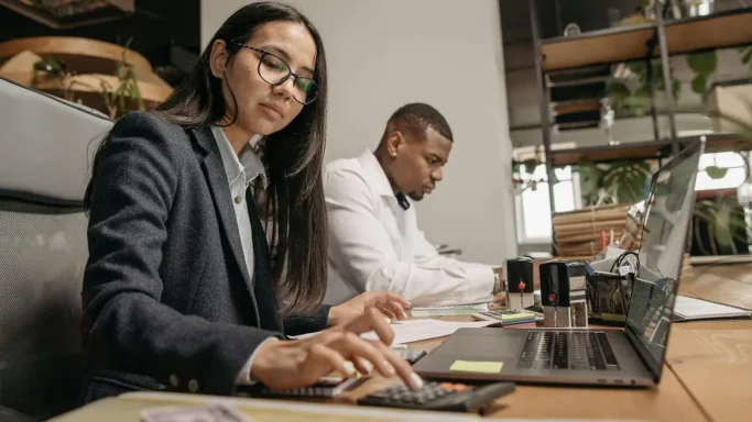 Woman using a calculator and laptop at a desk while a man works beside her.