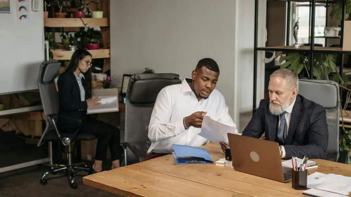 Two businessmen in an office reviewing documents together at a desk with a laptop, while a woman works in the background.