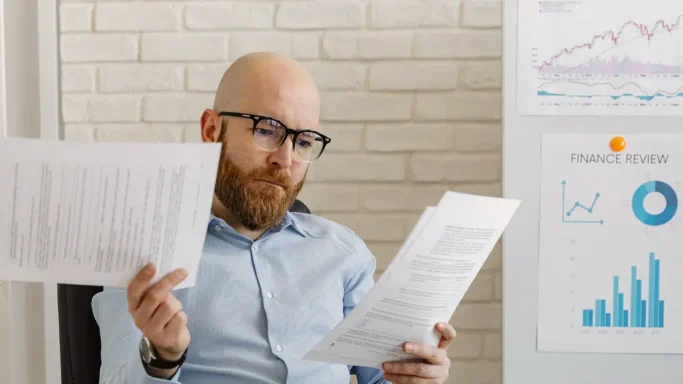 A man with glasses is reviewing documents while sitting near charts and graphs on the wall.