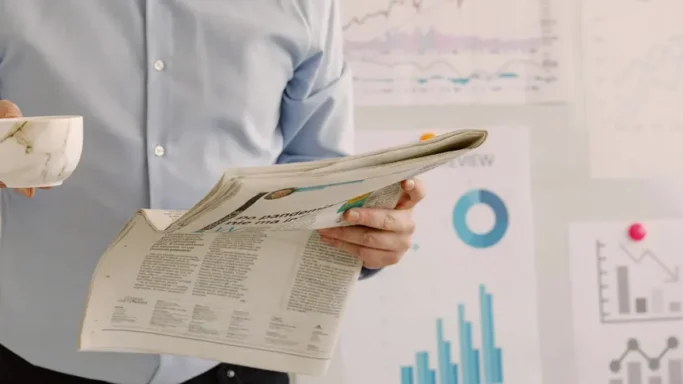 A person is holding a newspaper and a cup while standing near charts and graphs on the wall.
