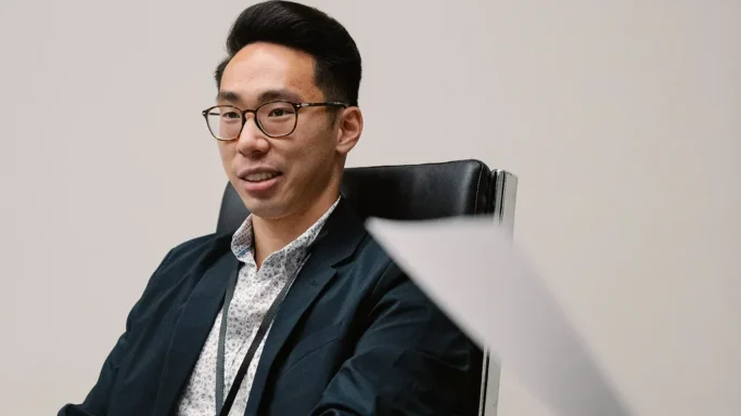A person wearing glasses and a blazer is sitting in a chair, smiling during a discussion.