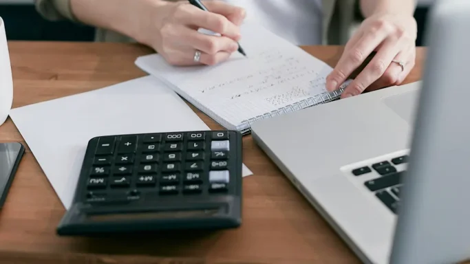 Person writing notes at a desk with a calculator and laptop.