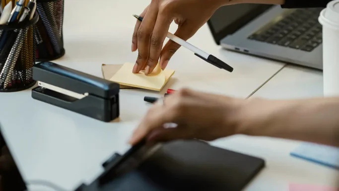 Close-up of hands working at a desk with notes, pen, and laptop.