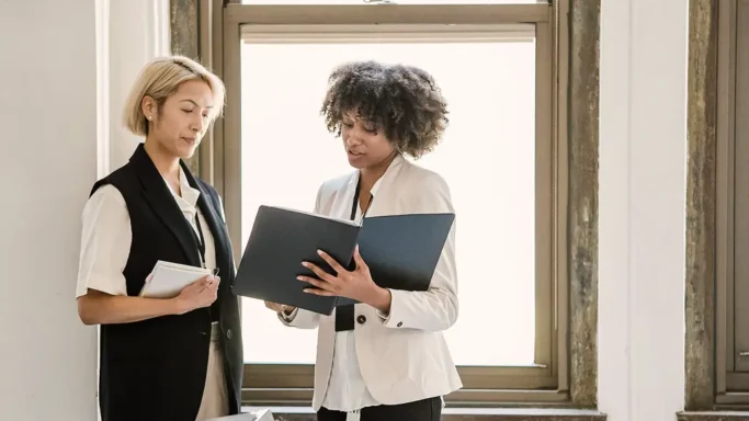 Two women standing by a window reviewing documents together.