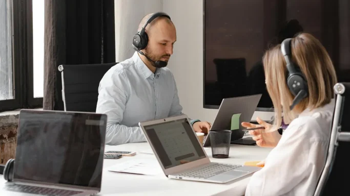 Two people wearing headsets are working on laptops during a support or conference call.