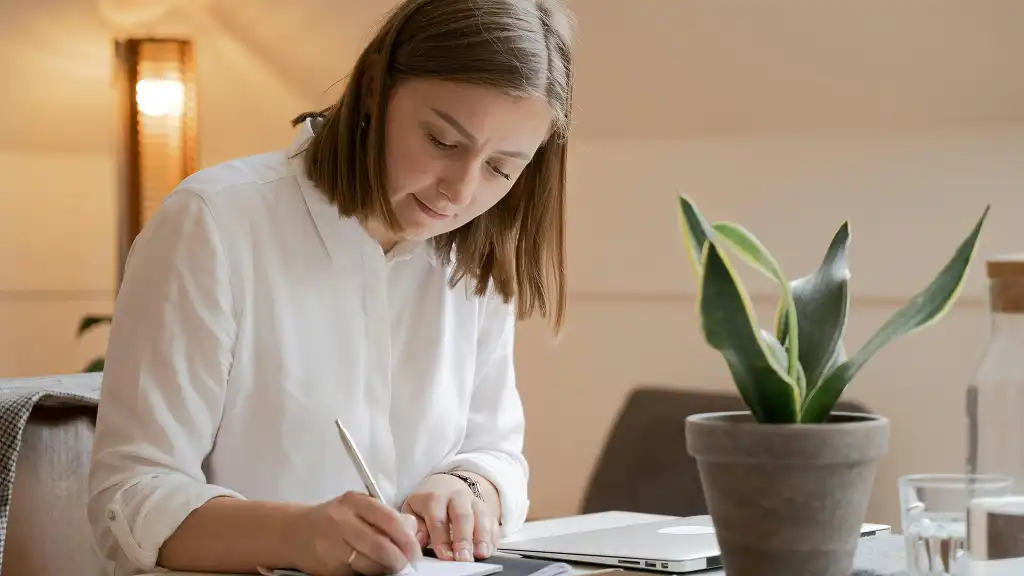 A woman in a white shirt focuses on writing at a table. Beside her is a laptop and a potted plant, creating a calm, productive atmosphere.