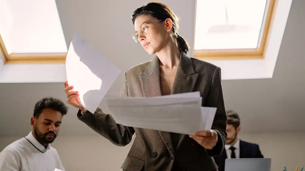 A focused woman in a brown blazer examines documents under soft skylight. Two colleagues work in the background, creating a professional atmosphere.