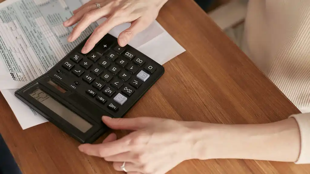 Hands use a large calculator on a wooden table, with a form and envelope nearby. The scene conveys focus and attention to financial tasks.