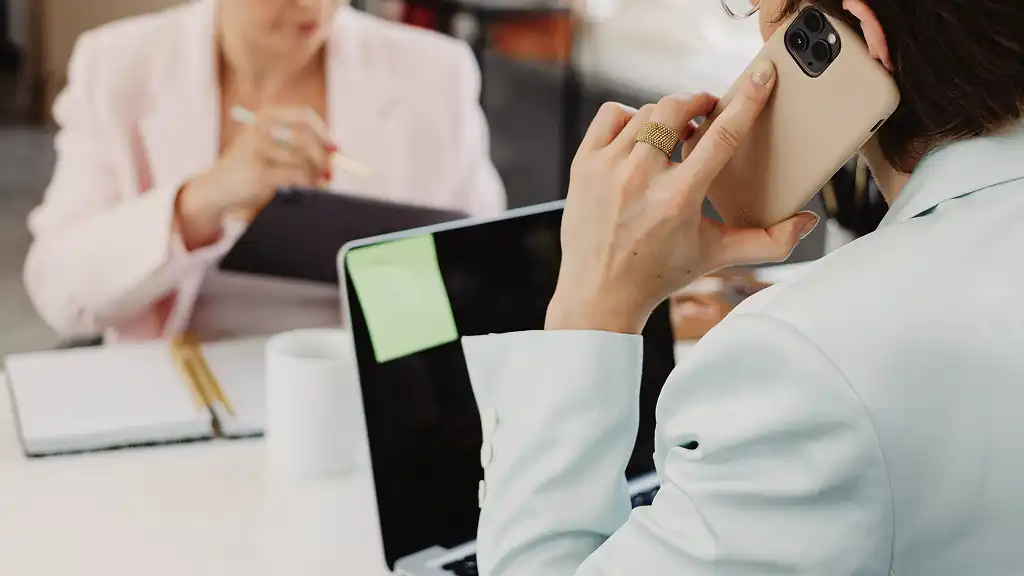 Two professionals in a meeting. One talks on a phone with a laptop open, a sticky note attached. The other writes on a tablet, focusing intently.
