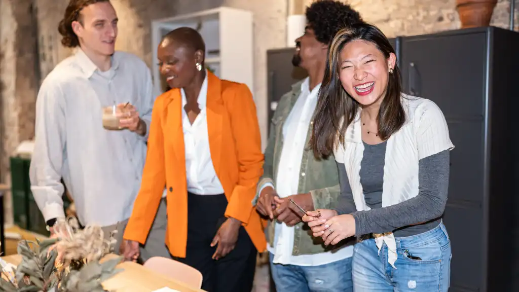 A diverse group of four people in casual attire share a joyful moment in an office setting. They are laughing, creating a warm and lively atmosphere.