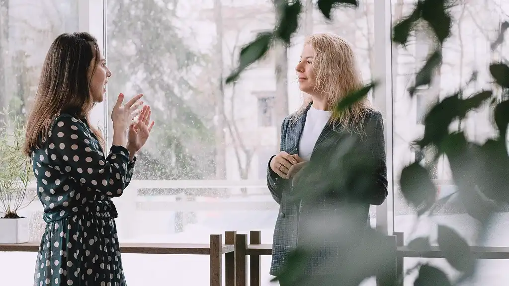 Two women engage in a lively conversation by a large window, surrounded by greenery. One gestures expressively. The atmosphere is casual and warm.