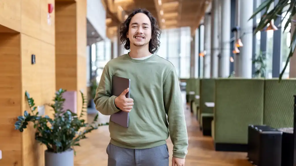 Smiling man in a green sweater holds a tablet in a modern, well-lit office with plants and green booths, conveying a relaxed, professional vibe.