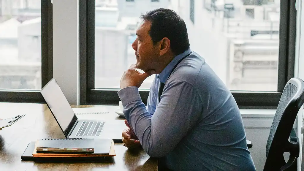 A man in a blue shirt sits at an office desk, focused on his laptop. The setting is a bright, modern office with large windows, conveying a professional and thoughtful atmosphere.