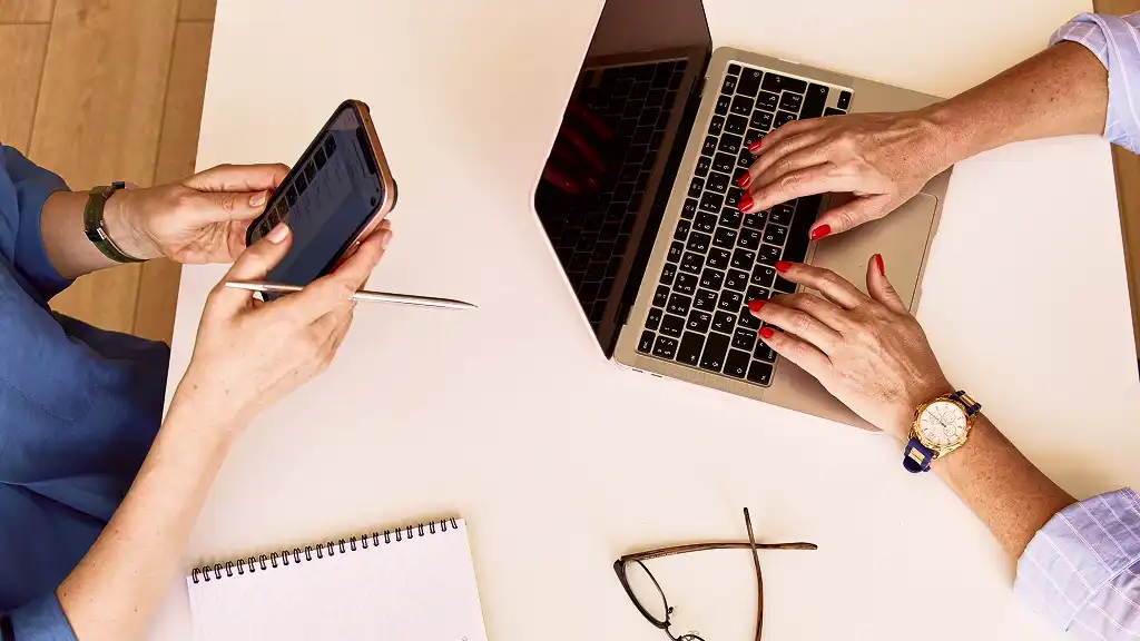 Two people working at a desk; one uses a smartphone, the other types on a laptop. Nearby are a notebook, pen, glasses, and a soft light conveys focus.