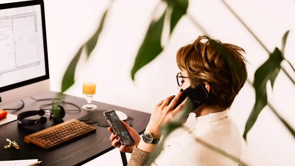 A woman with short hair talks on a phone while looking at her smartphone. She sits at a desk with a computer, keyboard, and headphones nearby. A plant is in the foreground, adding a natural element. The scene conveys a busy and focused office atmosphere.