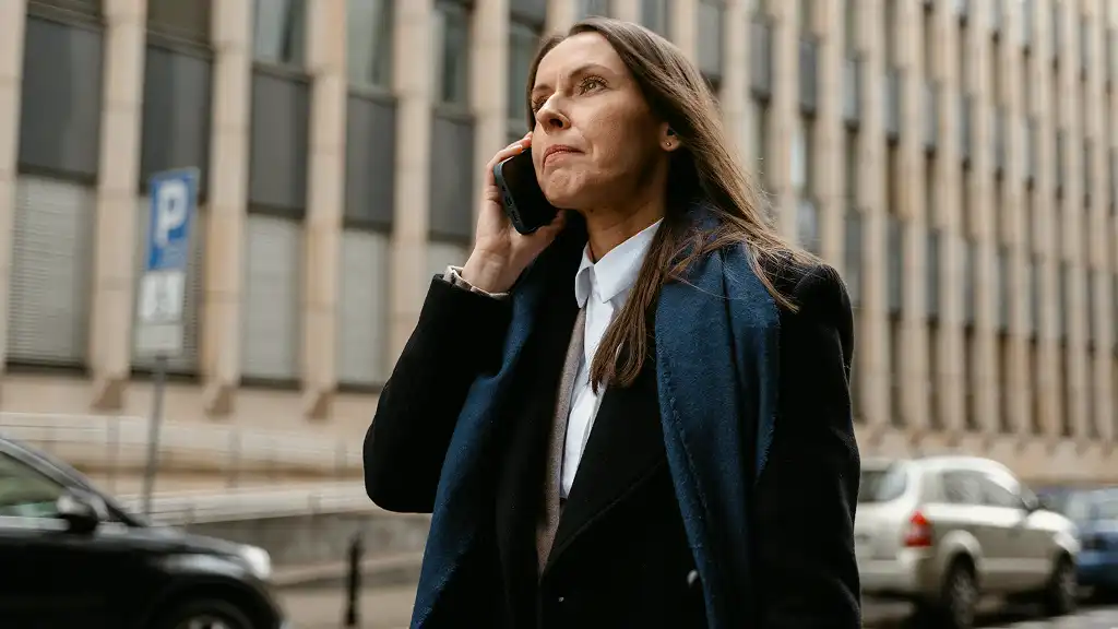 A woman in a dark coat walks down a city street, speaking on her phone, with a serious expression. Cars and a building with large windows are behind her.