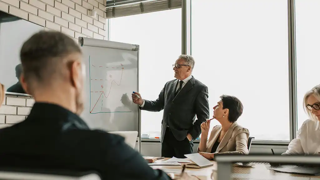 A man in a suit presents a graph on a flip chart in a bright conference room. Colleagues seated around a table focus attentively. Professional and engaged atmosphere.