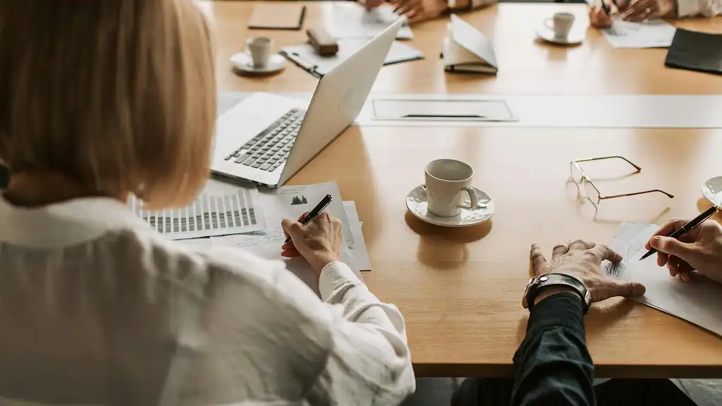 A group of people at a wooden table with laptops, documents, and coffee cups, engaged in a meeting. The atmosphere appears focused and collaborative.
