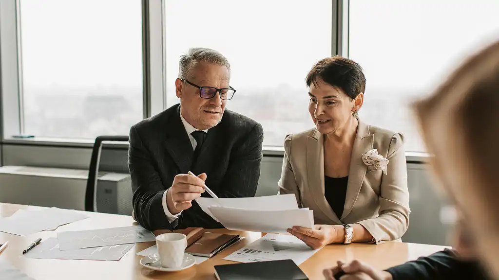 A man and a woman in business attire discuss documents at a conference table in a well-lit office, creating a professional and collaborative atmosphere.
