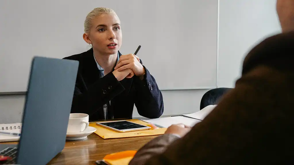 A person with short blonde hair, wearing a black jacket, attentively listens during a meeting. A laptop, tablet, and coffee cup are on the table. The setting is professional and focused.