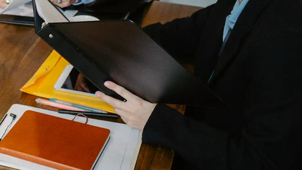 Person in a suit reading a black binder at a wooden desk with papers, a tablet, and a brown notebook. The setting is organized and professional.