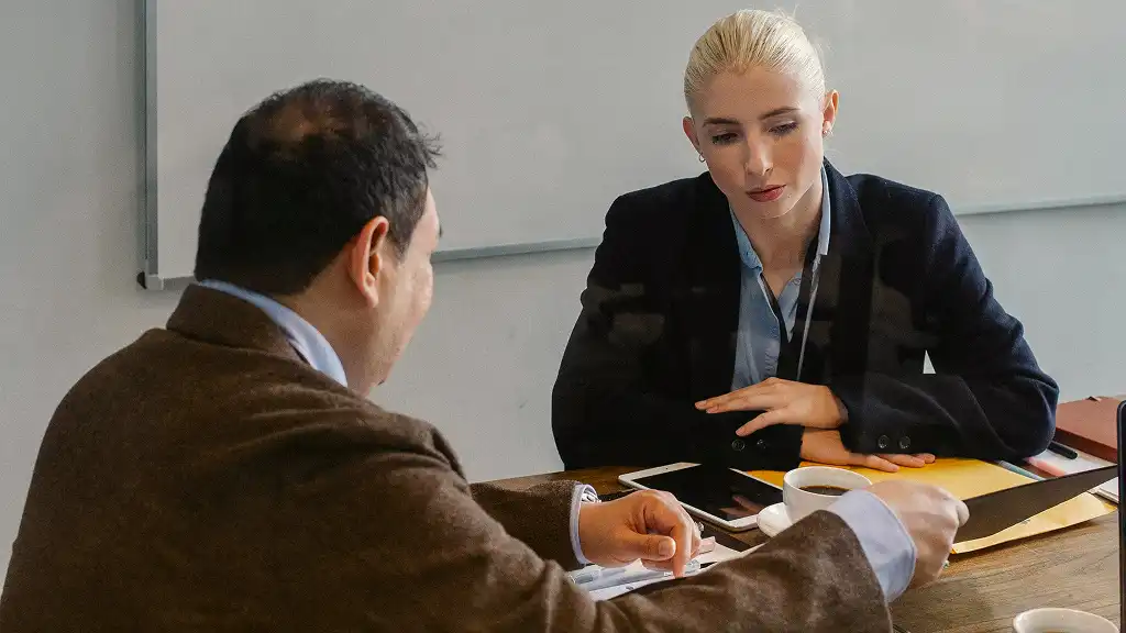 A man and a woman seated at a table in a professional setting, engaged in a serious discussion. The woman is attentive, and papers and a cup are on the table.