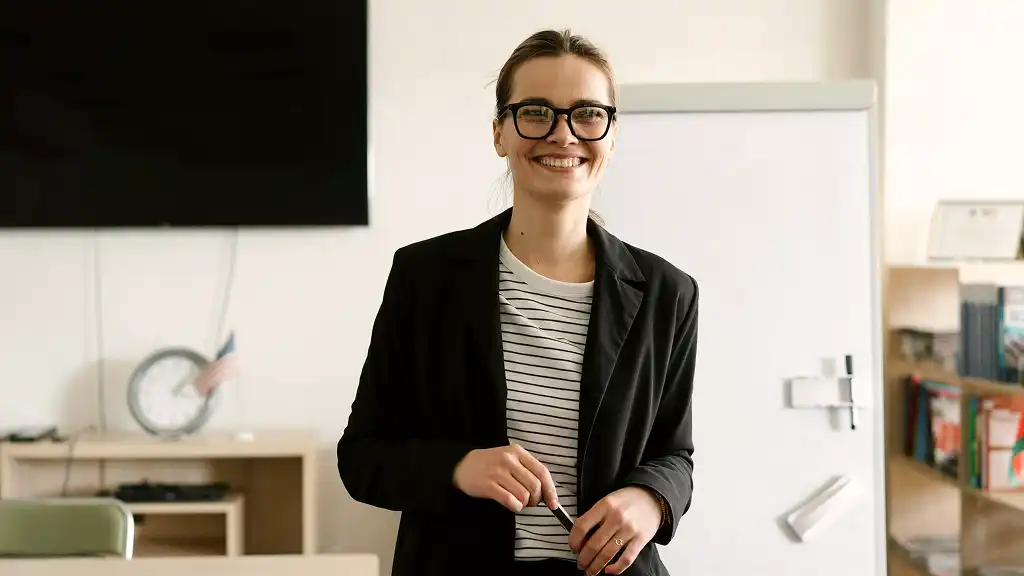 A woman in glasses and a black blazer smiles, standing in a classroom. She holds a marker. Behind her are a whiteboard and shelves with books, creating an educational atmosphere.