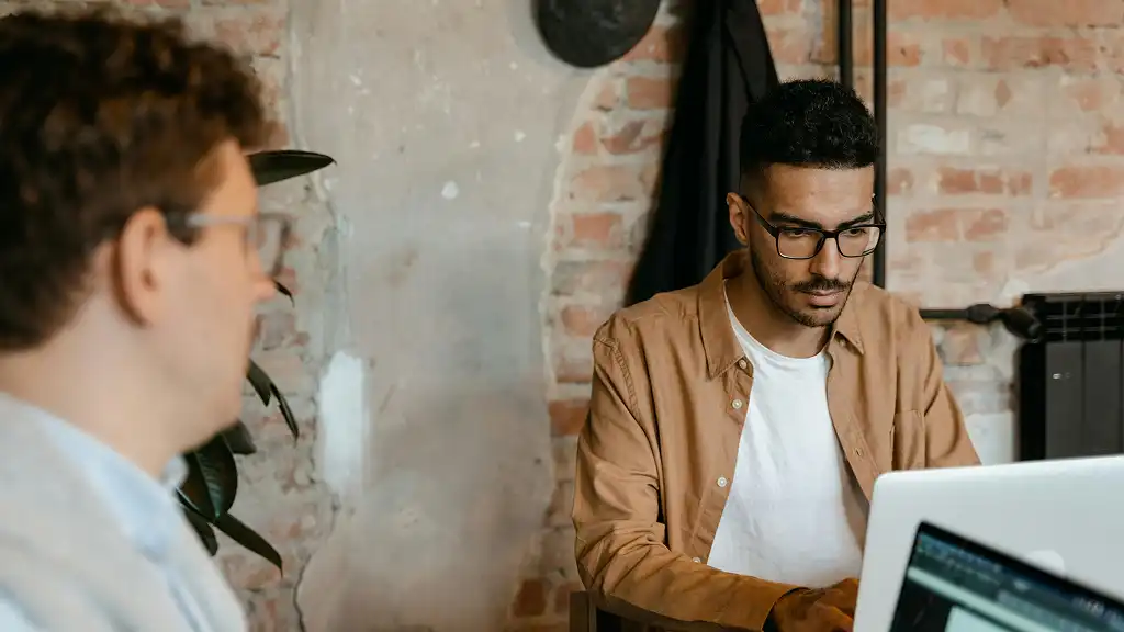Two men working on laptops in a brick-walled office. One, focused in the foreground, wears glasses and a brown shirt, projecting concentration.