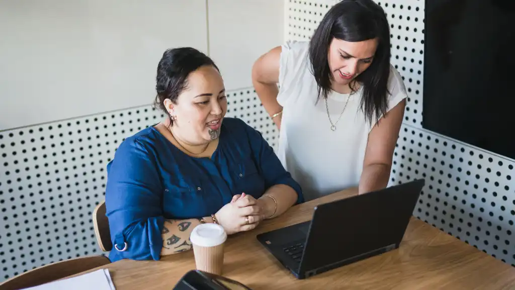 Two women collaborate at a wooden table, smiling as they look at a laptop. One wears a blue top, the other white. A coffee cup sits nearby, creating a warm, productive atmosphere.