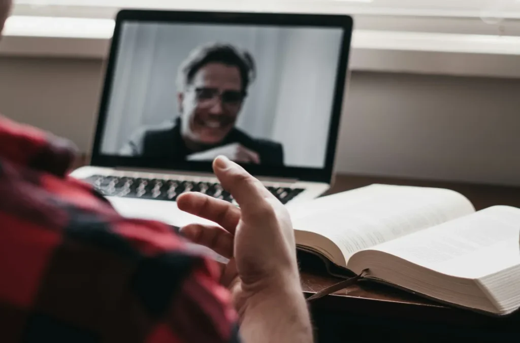 A person is on a video call using a laptop with an open book on the desk in front of them.