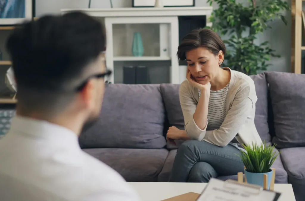 A woman sits on a couch looking distressed while talking to a therapist during a counseling session.