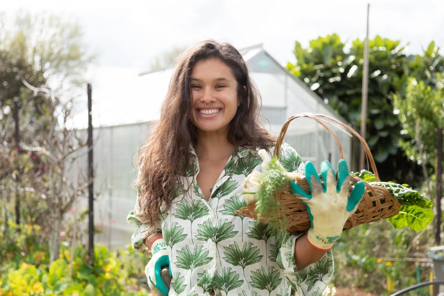 A smiling woman wearing gardening gloves and a green leaf-patterned shirt stands in a garden, holding a wicker basket filled with freshly picked greens and herbs, with a greenhouse and plants in the background.
