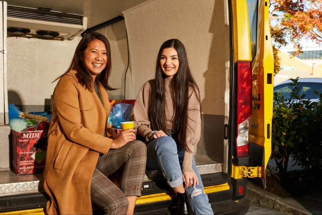 Two women seated in the back of a van, engaged in conversation and smiling at each other.