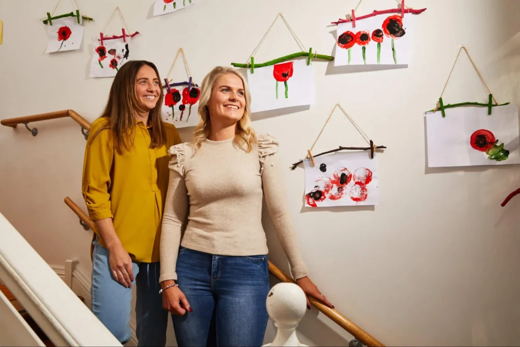 Two women stand on a stairway, surrounded by framed pictures hanging on the wall.