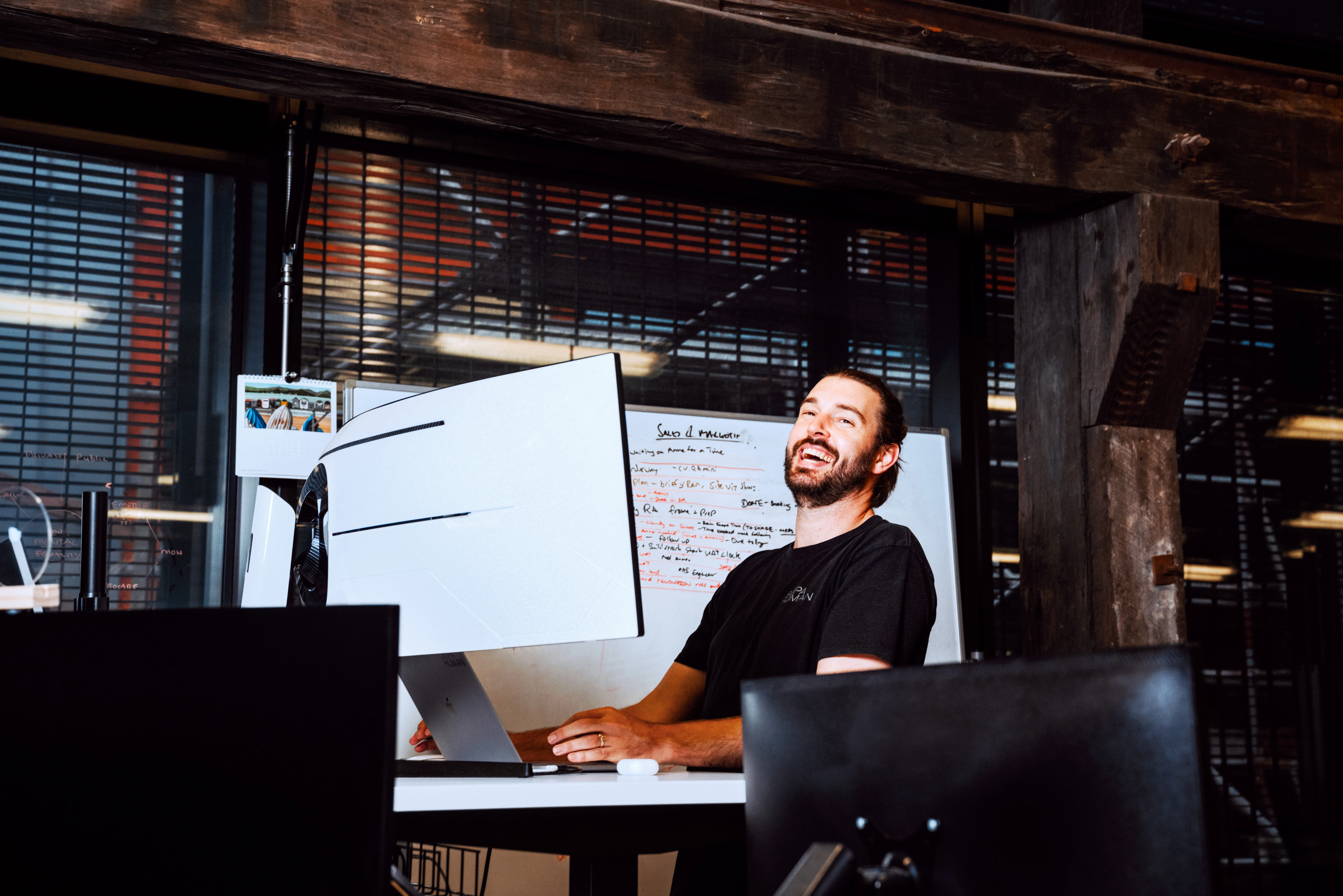 Man in a black shirt laughing while working at a desk with multiple monitors. Background features a whiteboard and industrial office setting. Joyful mood.