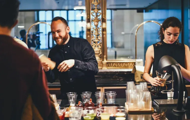 A smiling barista and a coworker preparing drinks behind a counter, highlighting the front-of-house staff employers manage and support in the hospitality industry.