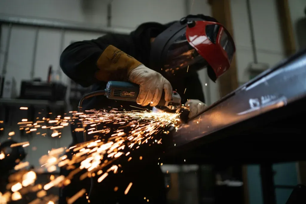 A worker in full safety gear grinding metal, creating sparks. AI in manufacturing can enhance site safety for employers through advanced predictive monitoring.