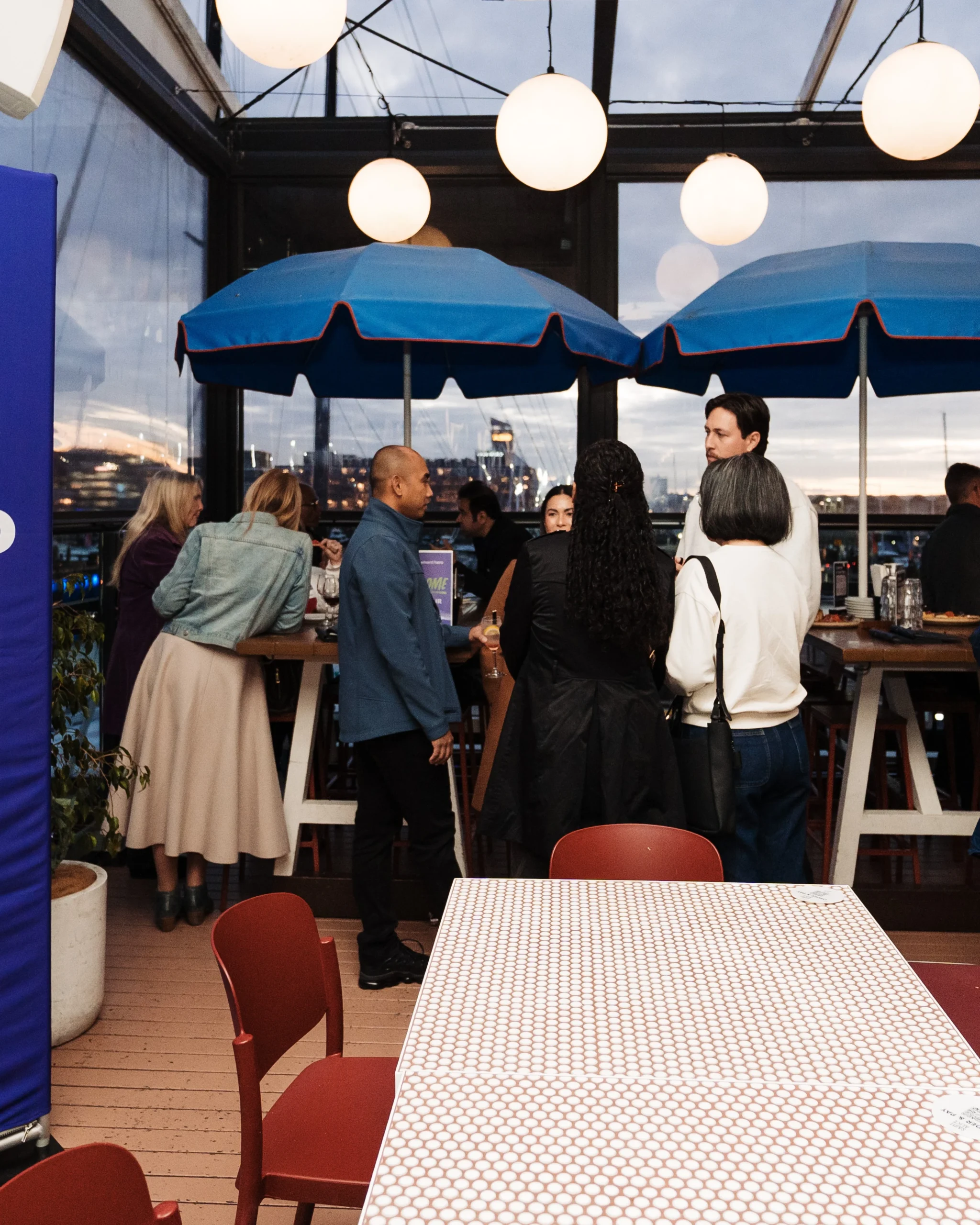 Guests socialize with drinks at a twilight networking event held on a glass-enclosed rooftop decorated with blue umbrellas and spherical pendant lamps.
