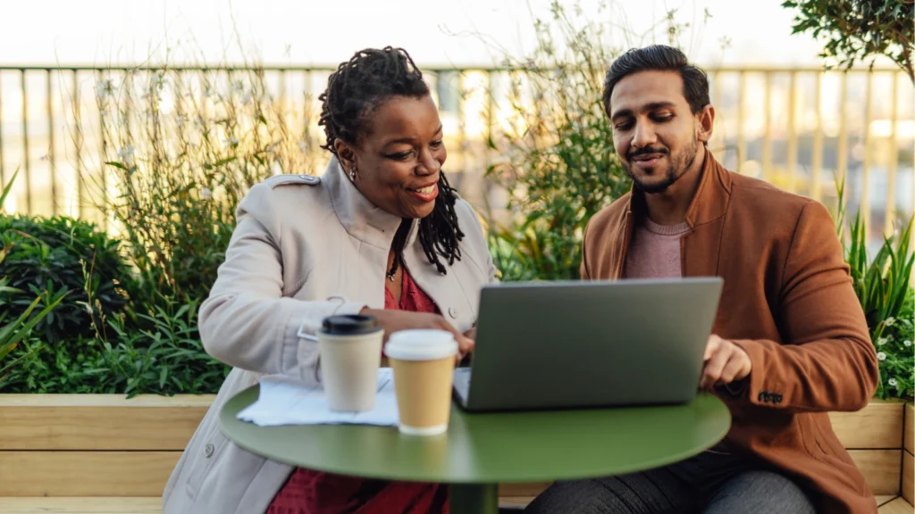 Two people sit at a green table outdoors, smiling and looking at a laptop. Coffee cups are on the table, surrounded by plants in a relaxed setting.