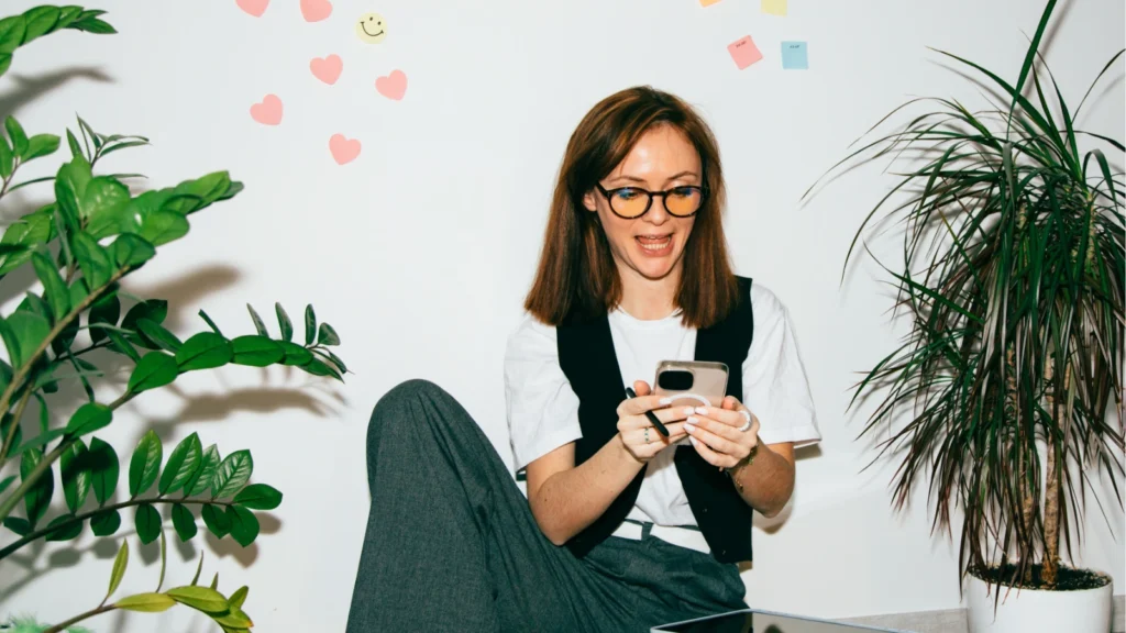 Woman with glasses smiling at phone, sitting on the floor with plants around. Pink heart notes and sticky notes on the wall create a cheerful vibe.