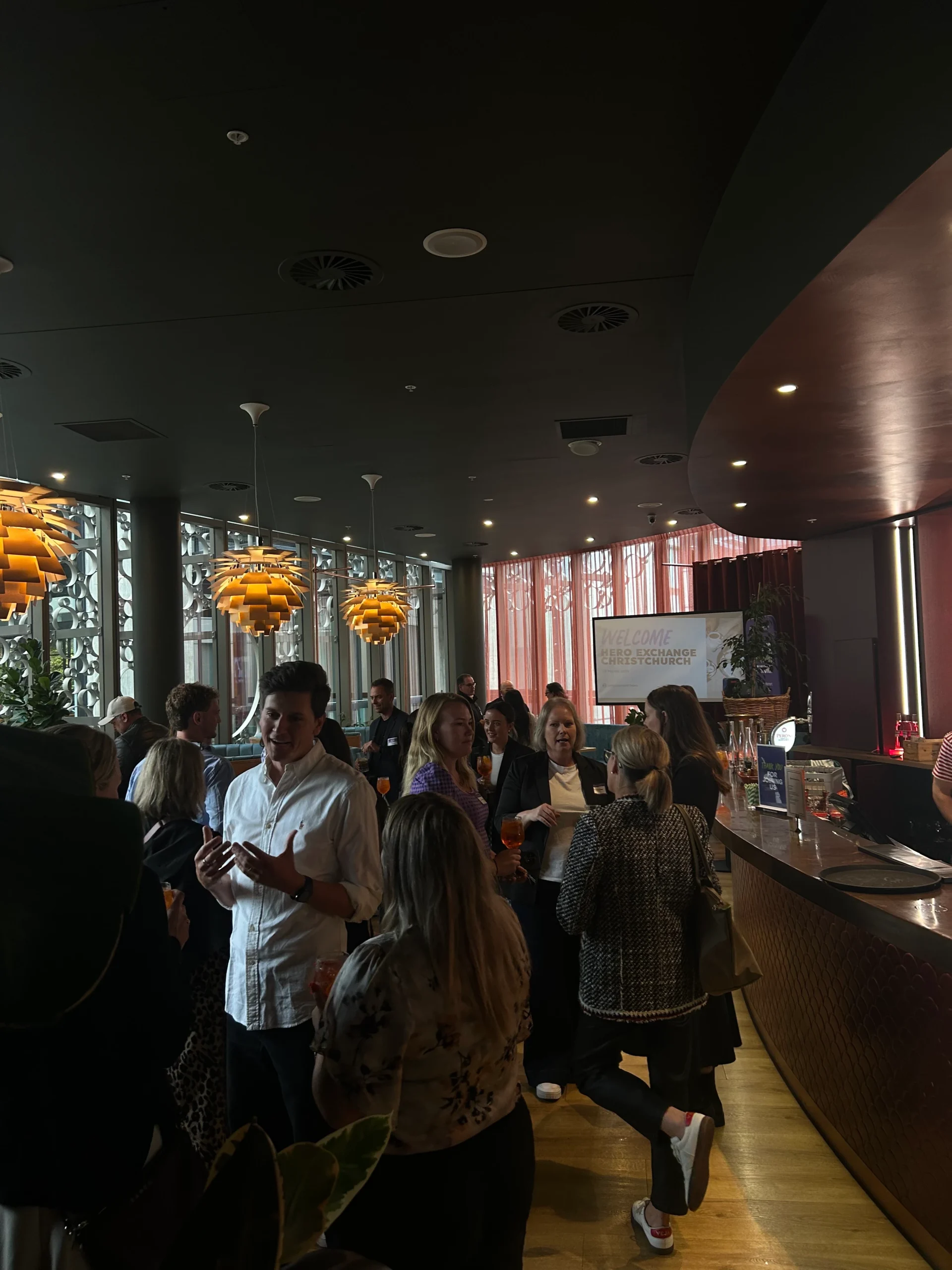 Attendees network in a stylish venue featuring a digital screen and distinctive tiered pendant lights.