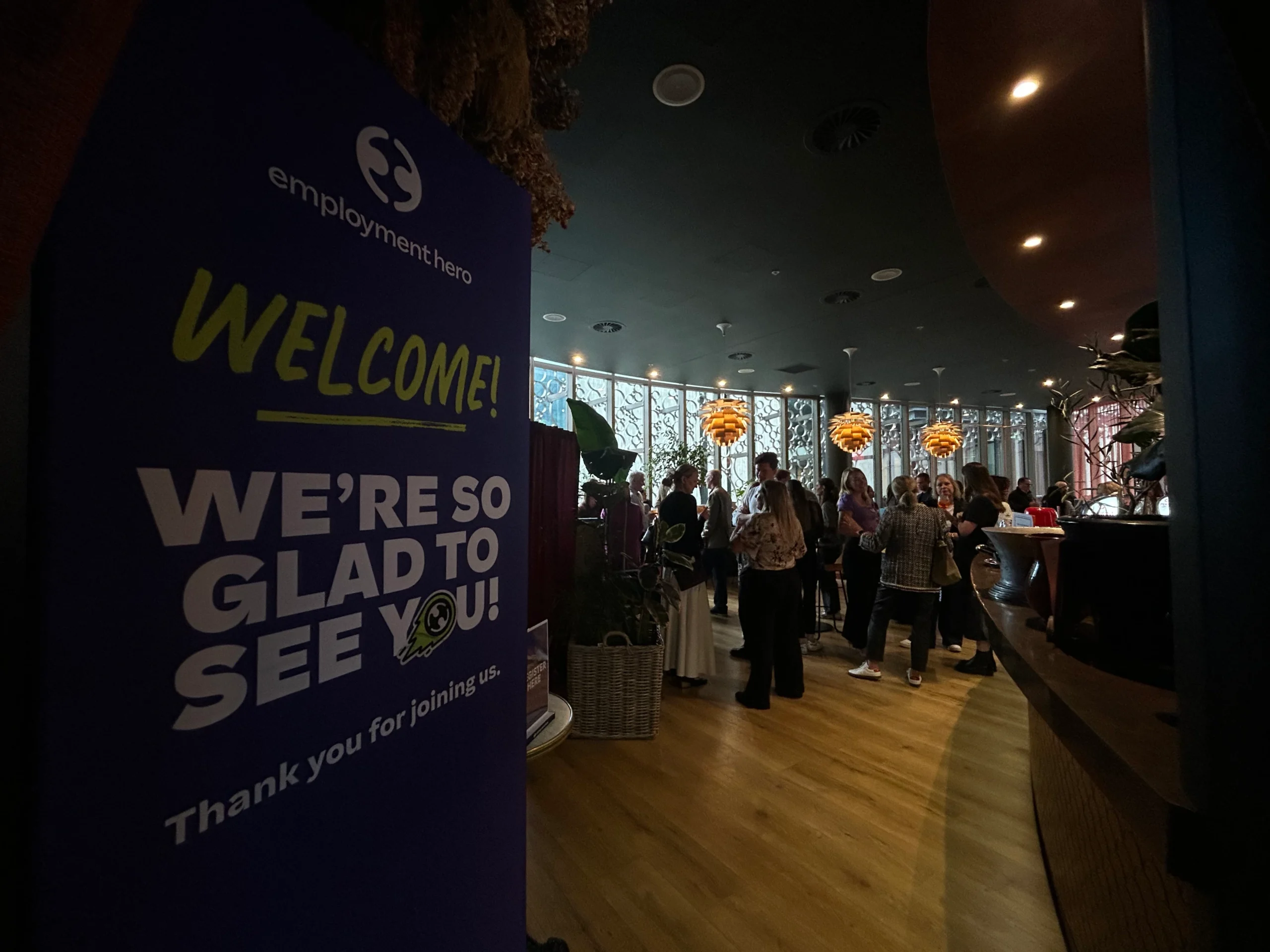 A tall blue welcome banner for an Employment Hero event stands in the foreground of a modern, dimly lit lounge where guests are socializing.