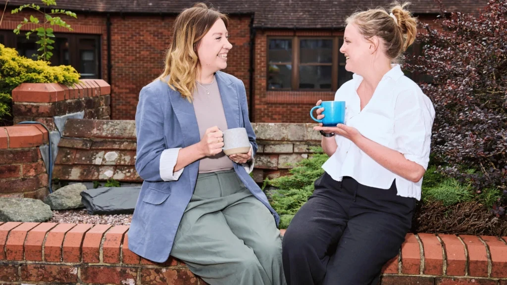 Two women seated on a brick wall, smiling at each other while holding mugs. They are surrounded by greenery and a brick building is in the background.
