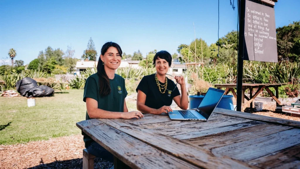 Two women smiling at a wooden table outdoors with a laptop. Behind them is a garden with trees, blue sky, and a sign. The mood is positive and collaborative.