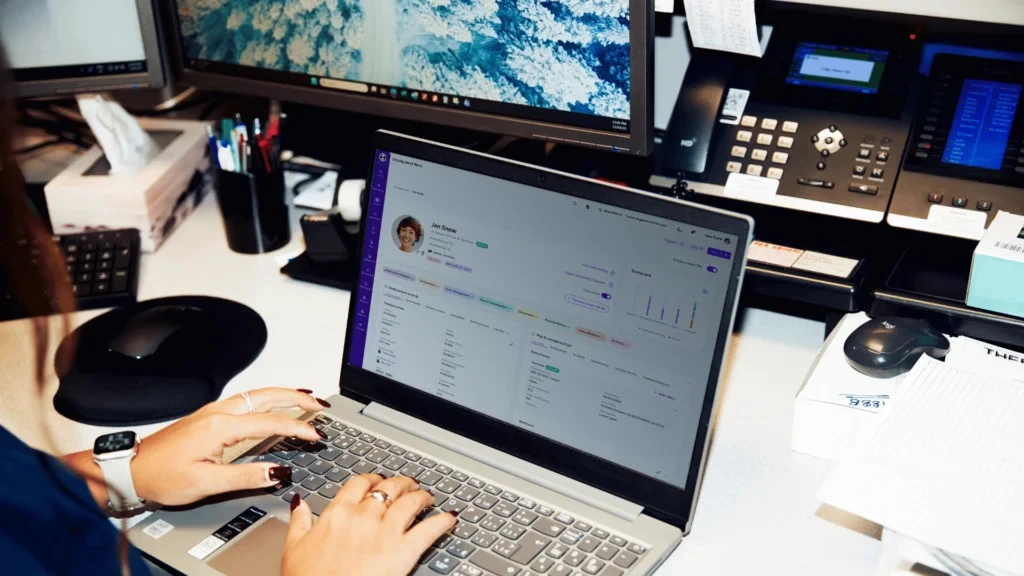 Person typing on a laptop in an office setting, with multiple screens displaying data. The atmosphere is focused, indicating a busy work environment.