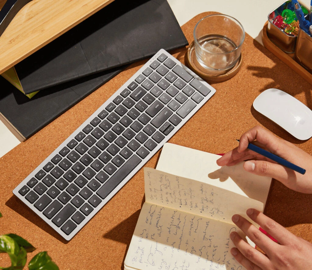 A tidy workspace with a wireless keyboard, mouse, and an open notebook with handwritten notes. A hand writes with a pencil, conveying focus.