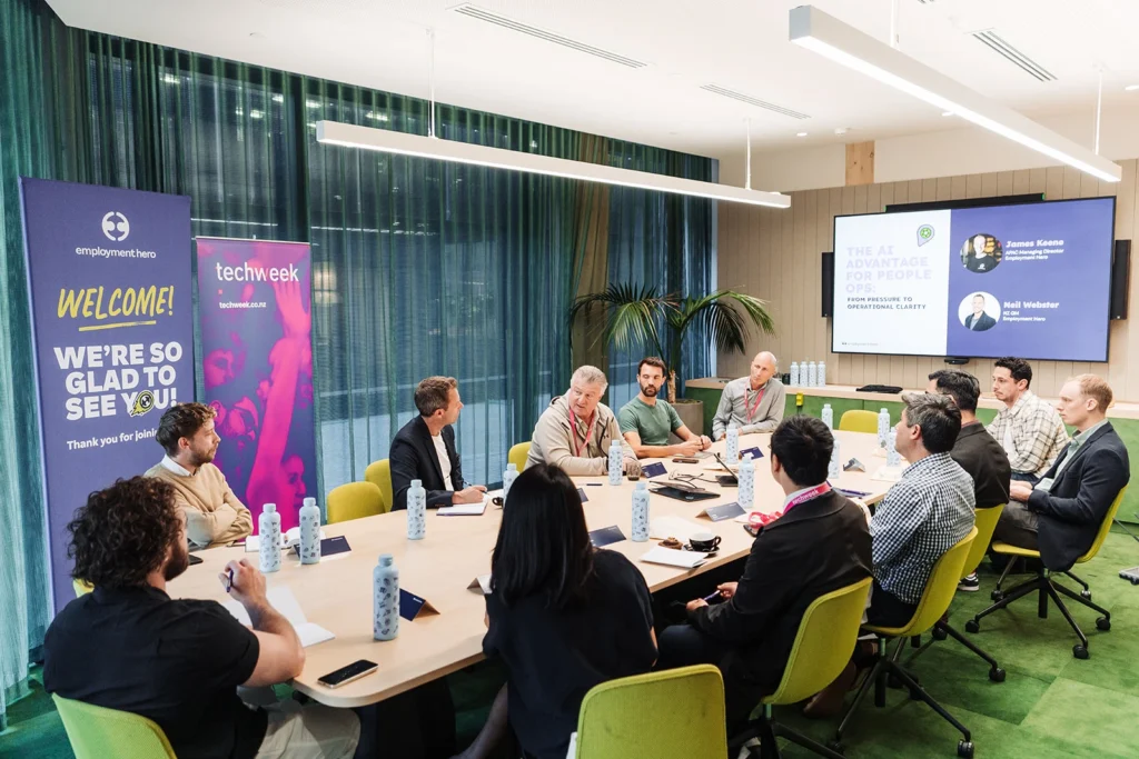 A group of people sit around a conference table in a modern meeting room. There are banners for Employment Hero and TechWeek. The mood is professional and collaborative.